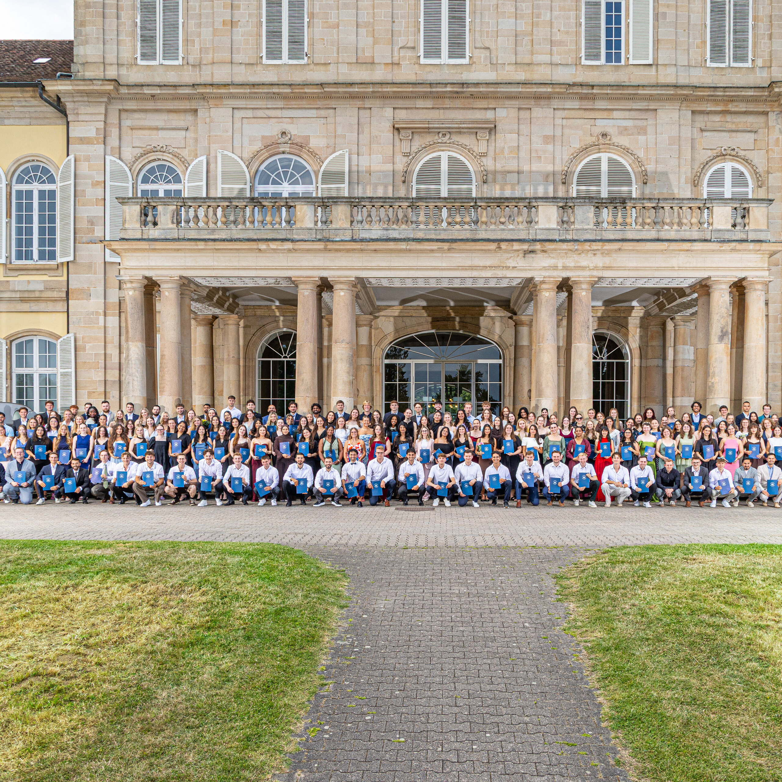 Foto: Eine große Gruppe von Personen steht vor dem Schloss Hohenheim. Das historische Gebäude mit symmetrischem Design und Säulen bildet den Hintergrund. Die Personen sind in mehreren Reihen aufgestellt.