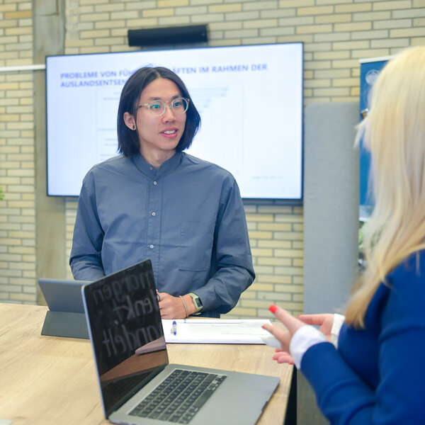 Foto: Drei Personen in einem modernen Büro, die an einem Tisch sitzen. Eine Person hält ein Tablet, während eine andere vor einem Laptop steht. Im Hintergrund sind Präsentationsfolien auf Bildschirmen sichtbar.