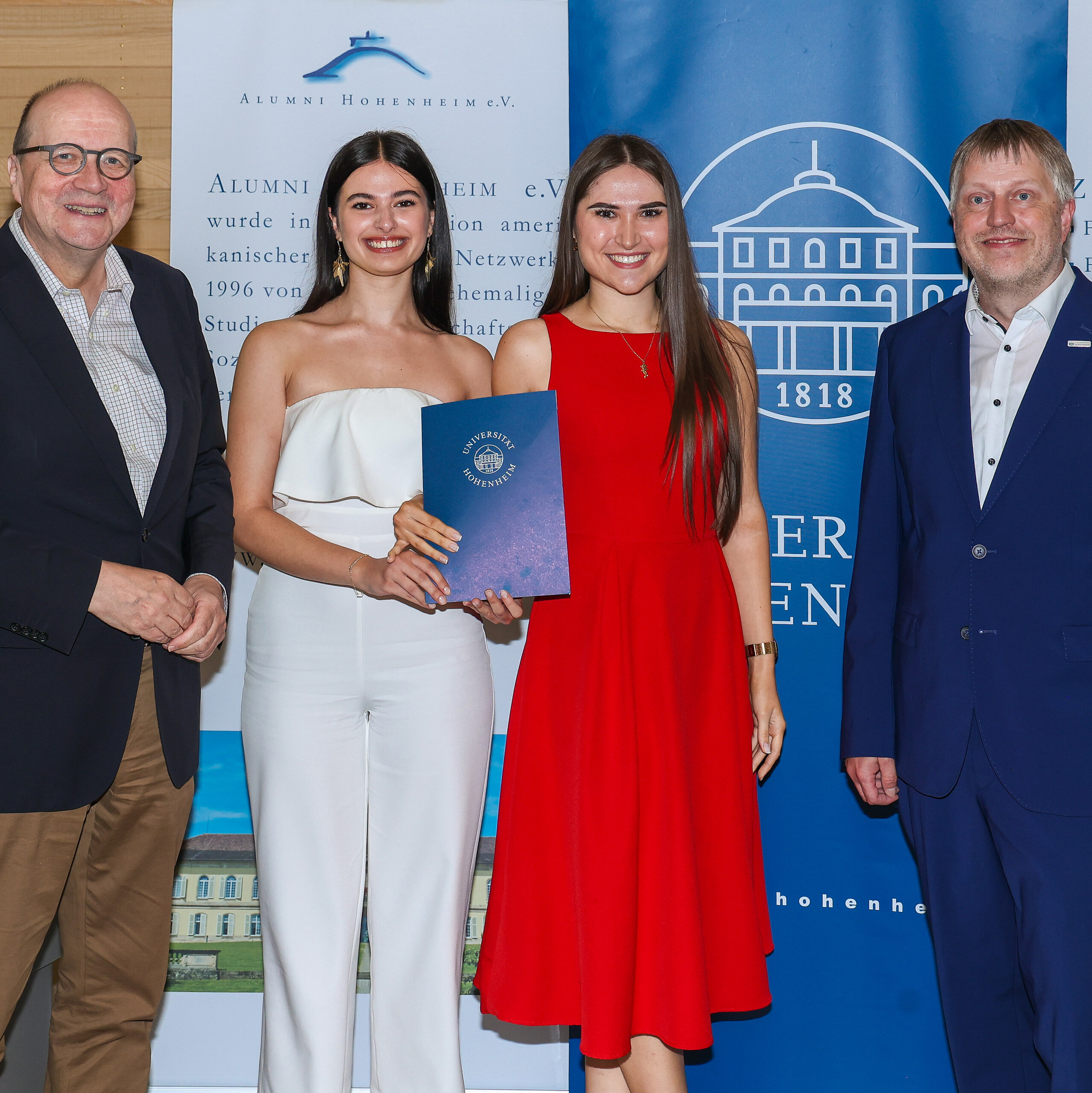 Foto: Vier Personen stehen vor einem Banner der Universität Hohenheim. Eine Person hält ein Zertifikat mit dem Logo der Universität. Im Hintergrund sind Banner mit Text über Alumni Hohenheim e.V. sichtbar.