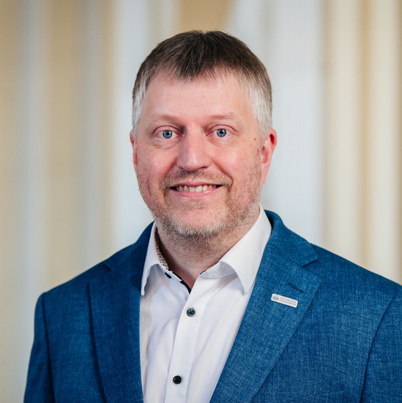 Foto: Eine Person trägt einen blauen Blazer und ein weißes Hemd vor einem neutralen Hintergrund.