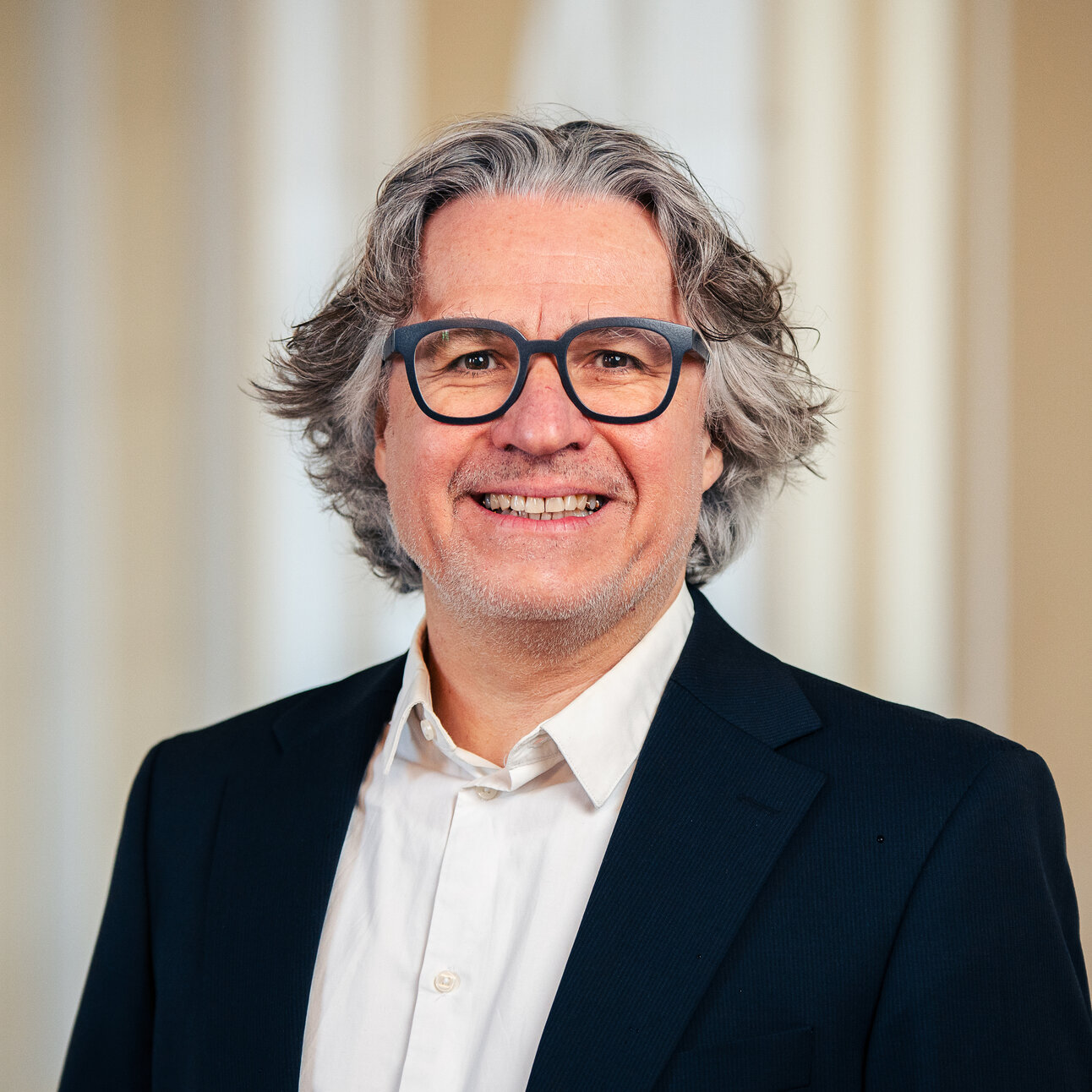 Foto: Eine Person in einem dunklen Anzug und weißem Hemd steht vor einem neutralen Hintergrund.