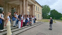 Foto: Eine Gruppe von Personen steht vor Schloss Hohenheim. Eine Person spricht zu der Gruppe. Einige Personen halten Taschen mit dem Logo der Universität Hohenheim. Das Gebäude im Hintergrund hat Säulen und große Fenster.