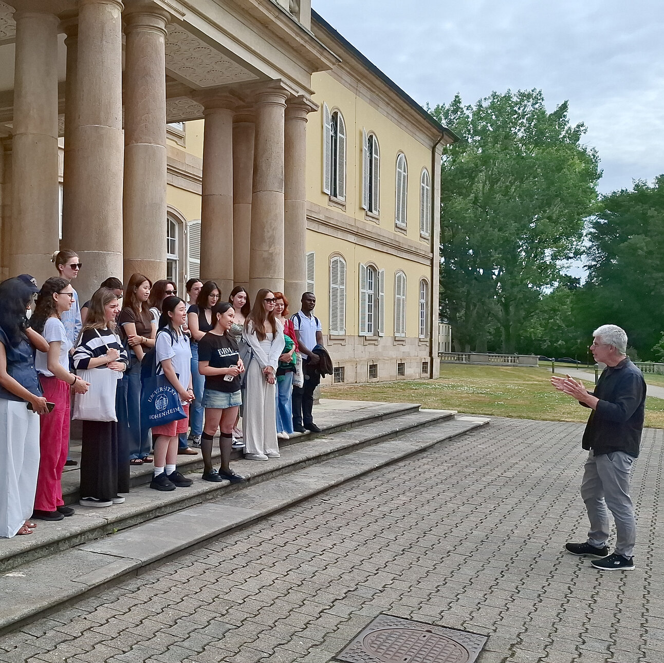Foto: Eine Gruppe von Personen steht vor Schloss Hohenheim. Eine Person spricht zu der Gruppe. Einige Personen halten Taschen mit dem Logo der Universität Hohenheim. Das Gebäude im Hintergrund hat Säulen und große Fenster.