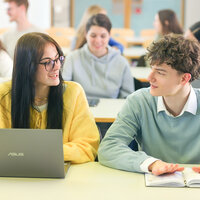Foto: Mehrere Personen sitzen in einem Hörsaal. Zwei Personen im Vordergrund unterhalten sich, eine mit einem Laptop, die andere mit einem Notizbuch. Im Hintergrund sind weitere Personen zu sehen.