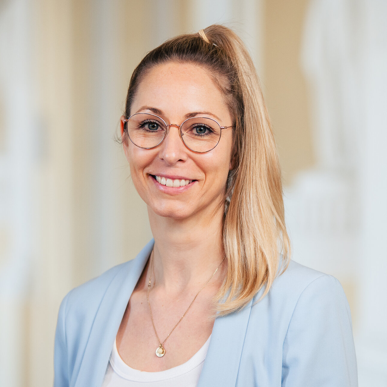 Foto: Eine Person trägt einen blauen Blazer und steht vor einem neutralen Hintergrund.