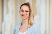 Foto: Eine Person trägt einen blauen Blazer und steht vor einem neutralen Hintergrund.