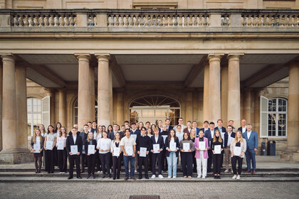 Foto: Eine Gruppe von Personen steht vor Schloss Hohenheim. Sie halten Zertifikate in den Händen und posieren auf den Stufen des Gebäudes mit Säulen und klassischer Architektur.
