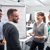Foto: Vier Personen stehen in einem Kursraum um ein Flipchart, das Diagramme zeigt. Eine Person hält einen Stift und spricht, während die anderen zuhören.