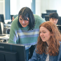 Foto: Mehrere Personen arbeiten in einem Computerraum. Eine Person steht und hilft anderen, die an Computern sitzen. Im Hintergrund sind weitere Computer zu sehen.