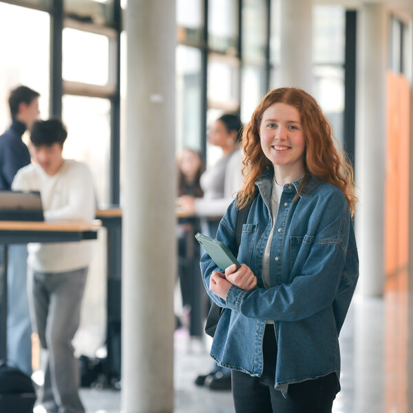 Foto: Eine Person in einer Jeansjacke steht in einem modernen Gebäude mit großen Fenstern. Im Hintergrund sind weitere Personen an einem Tisch mit Laptops zu sehen.