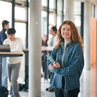 Foto: Eine Person in einer Jeansjacke steht in einem modernen Gebäude mit großen Fenstern. Im Hintergrund sind weitere Personen an einem Tisch mit Laptops zu sehen.
