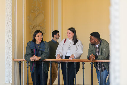 Foto: Eine Gruppe von Personen steht an einem Geländer im Schloss Hohenheim mit dekorativen Wänden im Hintergrund. Sie tragen verschiedene Jacken und Pullover und lehnen sich an ein Geländer.