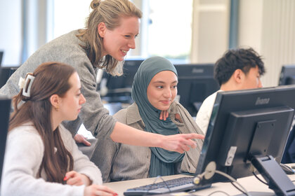 Foto: Mehrere Personen sitzen an Computern in einem Kursraum. Eine Person zeigt auf den Bildschirm eines Computers, während andere aufmerksam zuschauen. Die Umgebung wirkt modern und professionell.