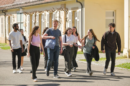 Foto: Eine Gruppe von Personen geht auf einem Gehweg vor einem Gebäude auf dem Campus der Universität Hohenheim. Eine Person trägt eine Tasche mit dem Logo der Universität.