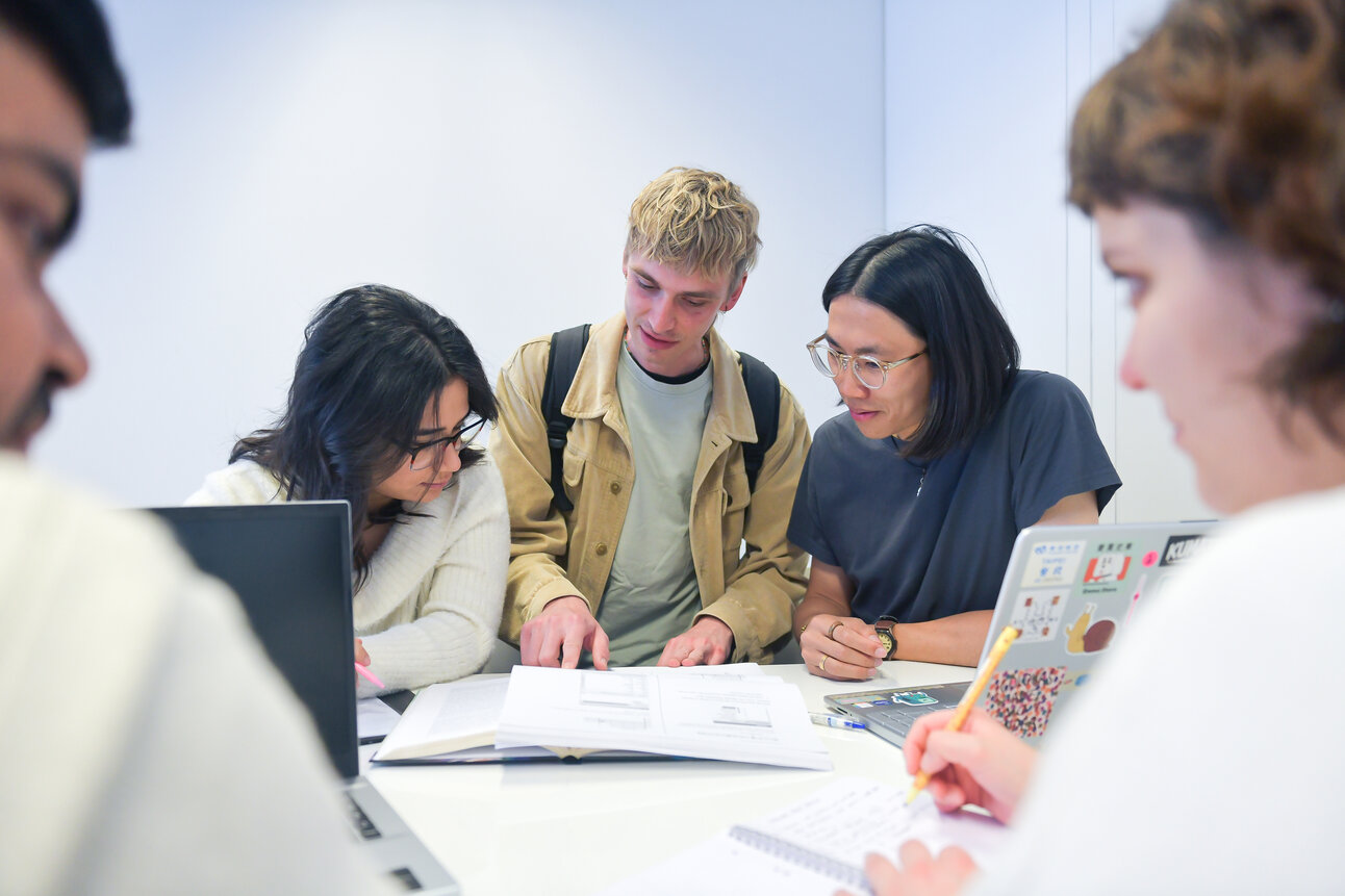 Foto: Eine Gruppe von Personen sitzt an einem Tisch und arbeitet zusammen. Auf dem Tisch liegen Bücher und Laptops. Eine Person macht Notizen auf einem Blatt Papier.