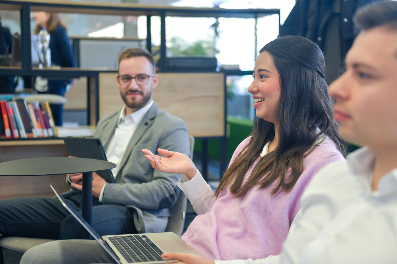 Foto: Drei Personen sitzen in einem modernen Büro. Eine Person hält einen Laptop, während eine andere ein Tablet hält. Im Hintergrund sind Bücherregale und Pflanzen sichtbar.