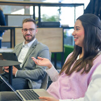 Foto: Drei Personen sitzen in einem modernen Büro. Eine Person hält einen Laptop, während eine andere ein Tablet hält. Im Hintergrund sind Bücherregale und Pflanzen sichtbar.