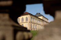 Foto: Schloss Hohenheim, ein großes historisches Gebäude mit symmetrischem Design und Kuppeldach, ist durch eine Öffnung in einem Steinzaun sichtbar.