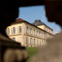 Foto: Schloss Hohenheim, ein großes historisches Gebäude mit symmetrischem Design und Kuppeldach, ist durch eine Öffnung in einem Steinzaun sichtbar.