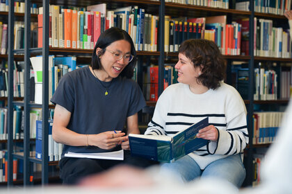 Foto: Zwei Personen sitzen in einer Bibliothek und betrachten gemeinsam ein Buch. Im Hintergrund sind Bücherregale mit verschiedenen Büchern sichtbar.