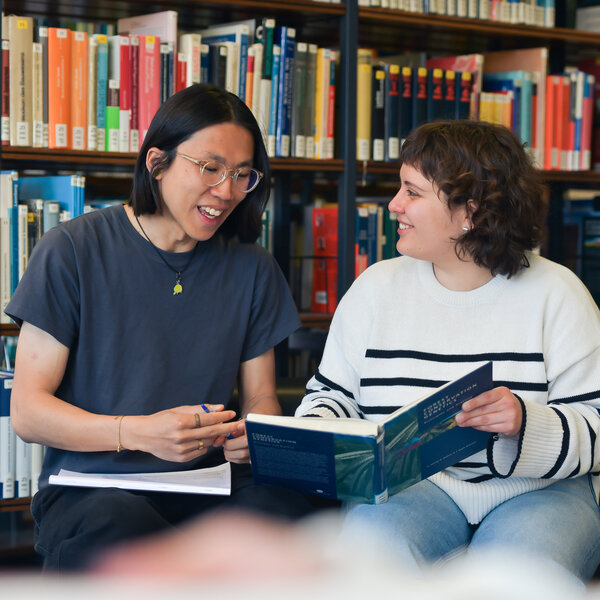 Foto: Zwei Personen sitzen in einer Bibliothek und betrachten gemeinsam ein Buch. Im Hintergrund sind Bücherregale mit verschiedenen Büchern sichtbar.