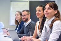 Foto: Vier Personen sitzen an einem Tisch in einem Konferenzraum. Eine Person benutzt einen Laptop, während die anderen Notizen machen. Im Hintergrund ist ein Whiteboard sichtbar.