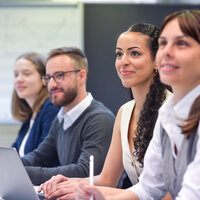 Foto: Vier Personen sitzen an einem Tisch in einem Konferenzraum. Eine Person benutzt einen Laptop, während die anderen Notizen machen. Im Hintergrund ist ein Whiteboard sichtbar.