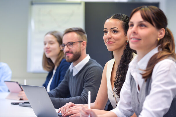 Foto: Vier Personen sitzen an einem Tisch in einem Konferenzraum. Eine Person benutzt einen Laptop, während die anderen Notizen machen. Im Hintergrund ist ein Whiteboard sichtbar.