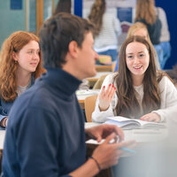 Foto: Mehrere Personen sitzen in einem Klassenzimmer und diskutieren. Eine Person hält ein Buch offen, während sie spricht. Im Hintergrund sind weitere Personen und eine Tafel sichtbar.