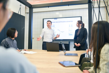Foto: Eine Person präsentiert vor einem Bildschirm mit Diagramm über Kapitalgesellschaften. Mehrere Personen sitzen an einem Tisch mit Laptops.