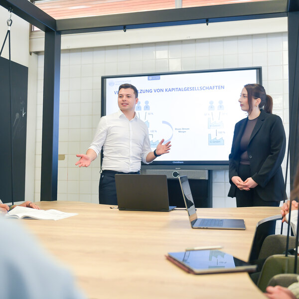 Foto: Eine Person präsentiert vor einem Bildschirm mit Diagramm über Kapitalgesellschaften. Mehrere Personen sitzen an einem Tisch mit Laptops.