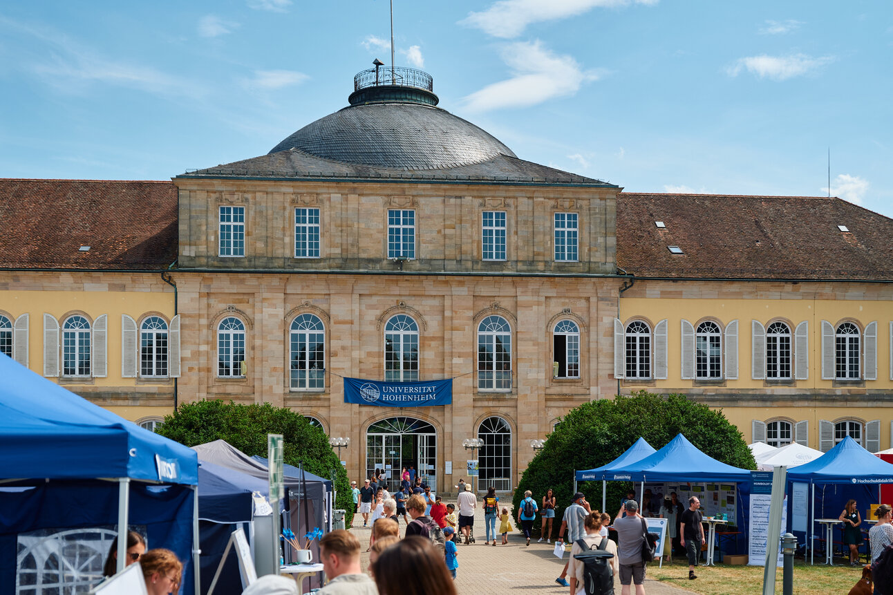 Foto: Schloss Hohenheim mit mehreren Personen, die an Informationsständen vorbeigehen. Ein Banner mit der Aufschrift 'Universität Hohenheim' ist sichtbar. Im Hintergrund ist das historische Gebäude mit Kuppeldach zu sehen.