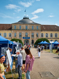 Foto: Schloss Hohenheim mit mehreren Personen, die an Informationsständen vorbeigehen. Ein Banner mit der Aufschrift 'Universität Hohenheim' ist sichtbar. Im Hintergrund ist das historische Gebäude mit Kuppeldach zu sehen.