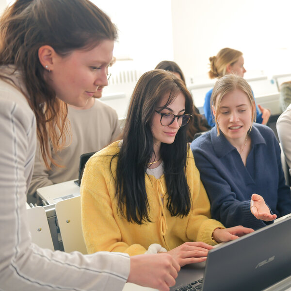 Foto: Eine Gruppe von Personen sitzt in einem Hörsaal und arbeitet gemeinsam an einem Laptop. Eine Person zeigt auf den Bildschirm, während andere aufmerksam zusehen.