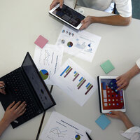 Foto: Drei Personen arbeiten an einem Tisch mit Laptops und Tablets. Auf dem Tisch liegen Diagramme und Notizen. Eine Person tippt auf einem Laptop, während die anderen Tablets verwenden.
