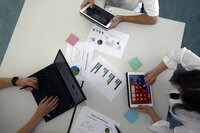 Foto: Drei Personen arbeiten an einem Tisch mit Laptops und Tablets. Auf dem Tisch liegen Diagramme und Notizen. Eine Person tippt auf einem Laptop, während die anderen Tablets verwenden.
