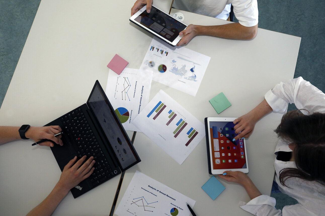 Foto: Drei Personen arbeiten an einem Tisch mit Laptops und Tablets. Auf dem Tisch liegen Diagramme und Notizen. Eine Person tippt auf einem Laptop, während die anderen Tablets verwenden.