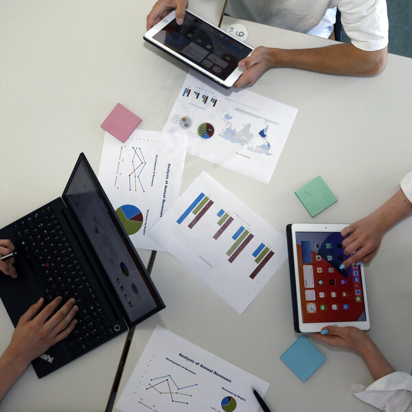 Foto: Drei Personen arbeiten an einem Tisch mit Laptops und Tablets. Auf dem Tisch liegen Diagramme und Notizen. Eine Person tippt auf einem Laptop, während die anderen Tablets verwenden.