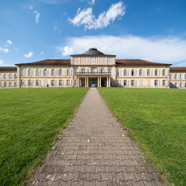 Foto: Schloss Hohenheim, ein großes historisches Gebäude mit symmetrischem Design und Kuppeldach, steht im Sonnenschein. Ein gepflasterter Weg führt durch einen gepflegten Rasen zum Eingang.