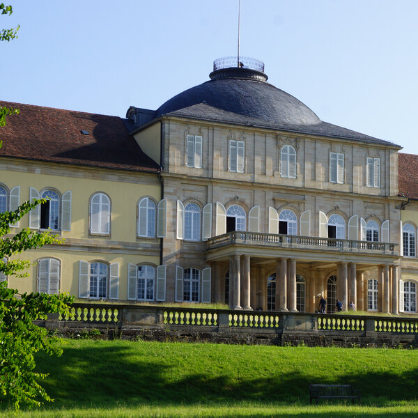 Foto: Schloss Hohenheim mit symmetrischem Design und Kuppeldach, umgeben von gepflegtem Rasen und Bäumen, bei sonnigem Wetter.