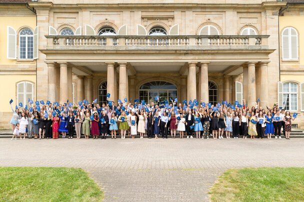 Foto: Eine große Gruppe von Personen steht vor Schloss Hohenheim. Sie halten blaue Mappen in die Luft, was auf eine Abschlussfeier hindeutet. Das Gebäude im Hintergrund hat klassische Architektur mit Säulen und großen Fenstern.