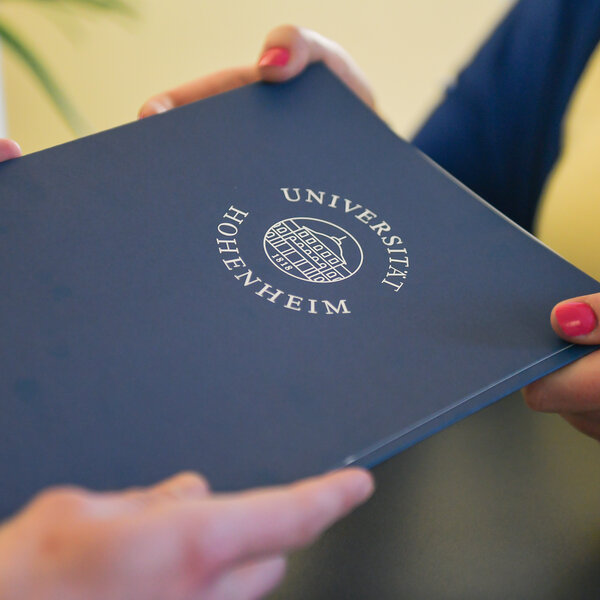 Foto: Zwei Personen halten ein blaues Dokument mit dem Logo der Universität Hohenheim. Das Logo zeigt ein Gebäude und den Schriftzug 'Universität Hohenheim'.