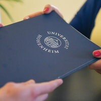 Foto: Zwei Personen halten ein blaues Dokument mit dem Logo der Universität Hohenheim. Das Logo zeigt ein Gebäude und den Schriftzug 'Universität Hohenheim'.