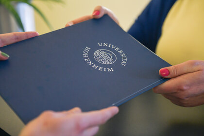 Foto: Zwei Personen halten ein blaues Dokument mit dem Logo der Universität Hohenheim. Das Logo zeigt ein Gebäude und den Schriftzug 'Universität Hohenheim'.