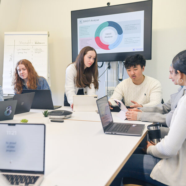 Foto: Eine Gruppe von Personen sitzt an einem Tisch mit Laptops. Ein Bildschirm zeigt eine SWOT-Analyse. Ein Flipchart im Hintergrund enthält die Wörter 'Strengths', 'Weaknesses', 'Opportunities' und 'Threats'.