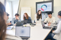 Foto: Eine Gruppe von Personen sitzt an einem Tisch mit Laptops. Ein Bildschirm zeigt eine SWOT-Analyse. Ein Flipchart im Hintergrund enthält die Wörter 'Strengths', 'Weaknesses', 'Opportunities' und 'Threats'.