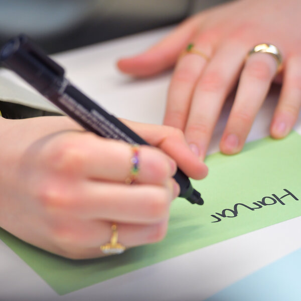 Foto: Eine Person schreibt mit einem schwarzen Stift auf eine grüne Karteikarte. Der Text auf der Karte lautet 'Honour'. Auf dem Tisch liegen weitere bunte Karteikarten und ein Tablet.