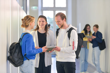 Foto: Drei Personen stehen in einem Flur und unterhalten sich. Eine Person hält ein Tablet. Im Hintergrund sind zwei weitere Personen zu sehen, die ebenfalls ein Tablet betrachten.