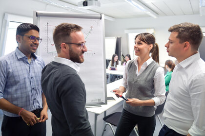 Foto: Vier Personen stehen in einem Kursraum um ein Flipchart, das Diagramme zeigt. Eine Person hält einen Stift und spricht, während die anderen zuhören.
