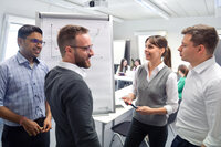 Foto: Vier Personen stehen in einem Kursraum um ein Flipchart, das Diagramme zeigt. Eine Person hält einen Stift und spricht, während die anderen zuhören.